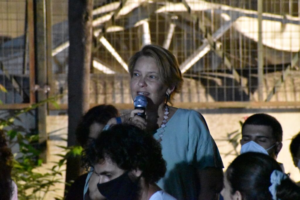 Η Μαίρη Χιόνη μιλά όρθια από το κοινό με φορητό μικρόφωνο, έχει ανοιχτόχρωμα κοντά μαλλιά, φορά ανοιχτόχρωμη μπλούζα και μακρύ κολιέ.
