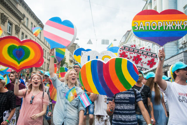 Στιγμιότυπο από την πορεία του Pride στο Κίεβο το 2019. Άτομα φωνάζοντας συνθήματα και χαμογελώντας κρατούν πικέτες σε σχήμα καρδιάς στα χρώματα του ουράνιου τόξου και της τρανς σημαίας (ροζ/γαλάζιο).